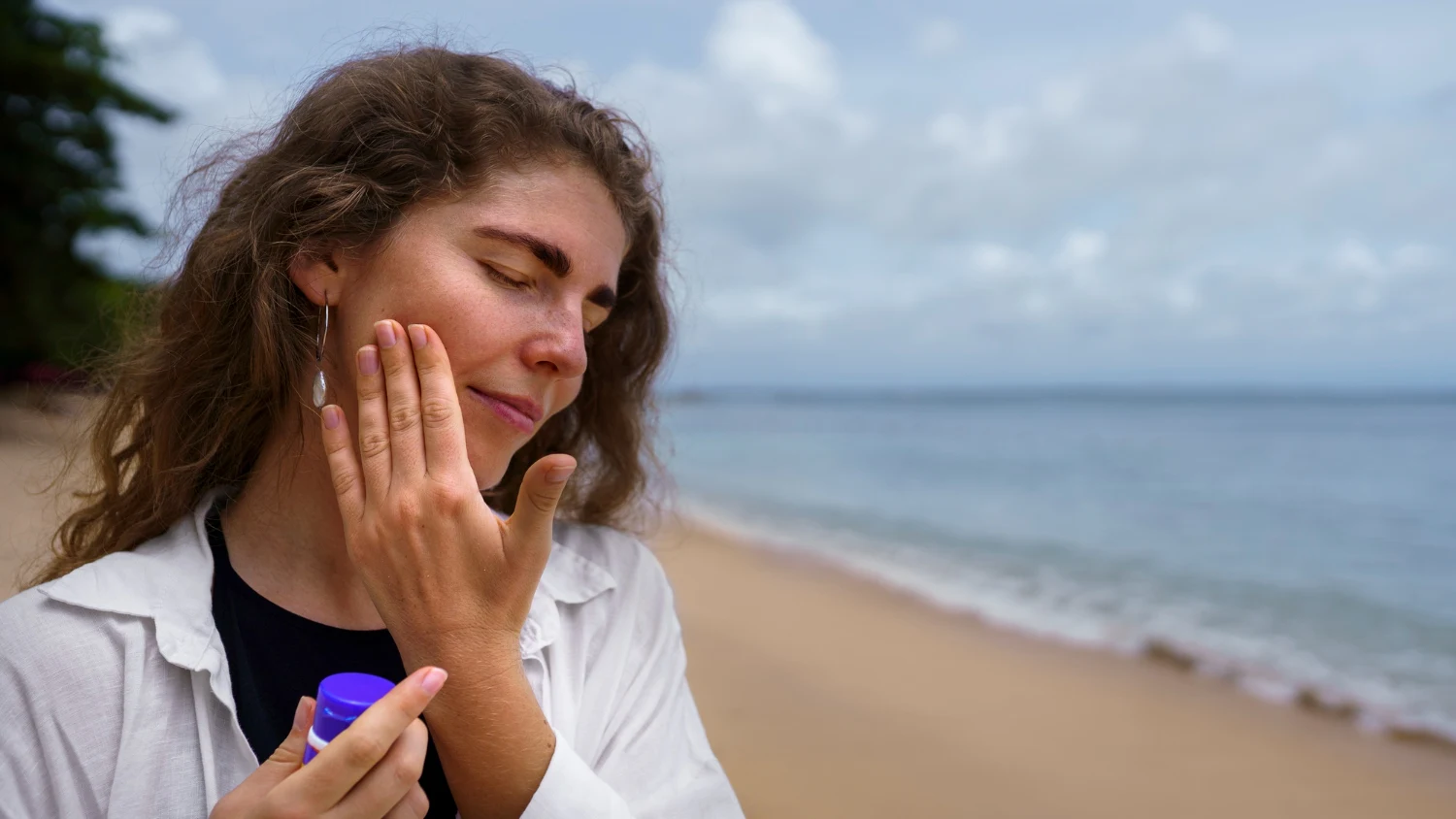 Woman applying sunscreen to her cheek at the beach, highlighting sun protection as part of a skincare routine for combination skin in humid weather.